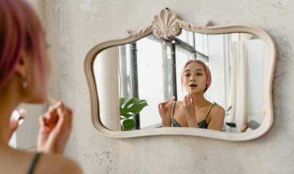 Young woman with pink hair applying makeup in front of ornate mirror indoors, embracing self-care.